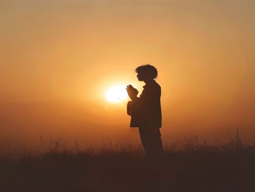 A silhouette of a person standing in prayer with hands slightly raised, captured against the soft, warm light of a sunri